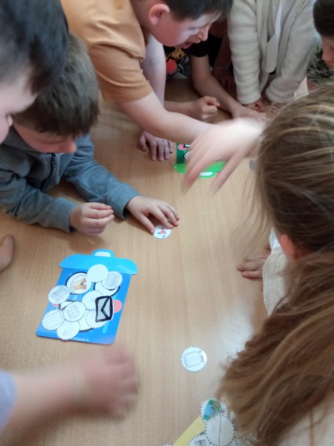 Dzieci pochylone nad stołem, na którym rozłożone są kolorowe żetony z rysunkami przedmiotów. Dzieci dopasowują żetony do odpowiedniego koloru kosza na śmieci.