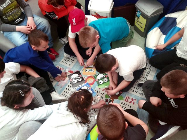 Dzieci pochylone nad dywanem, na którym leżą kolorowe rysunki koszy na śmieci zgodne z zasadami segregacji.